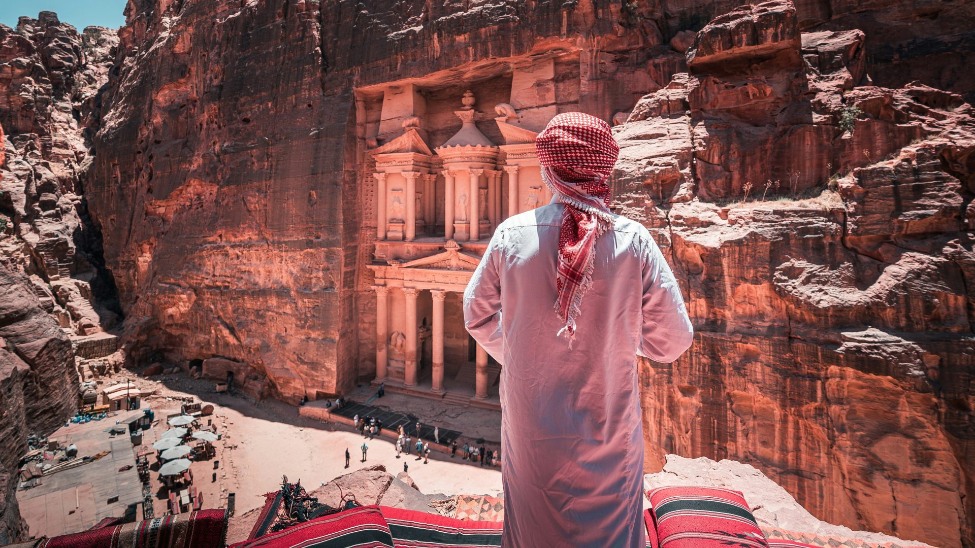 Bedouin man wearing traditional Jordanian attire overlooking Al-Khazneh (The Treasury) in Petra, Jordan, surrounded by dramatic rose-red sandstone cliffs