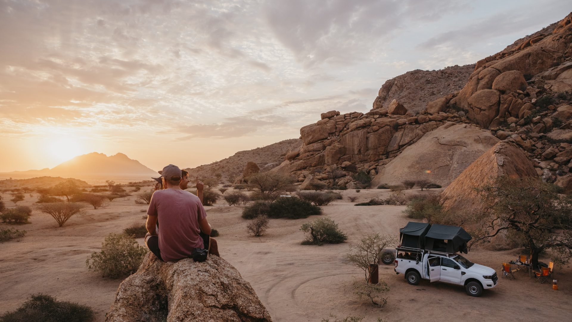 Tourist sitting on a rock overlooking a desert landscape at sunset, with an off-road 4x4 jeep nearby, adventure travel and desert safari experience