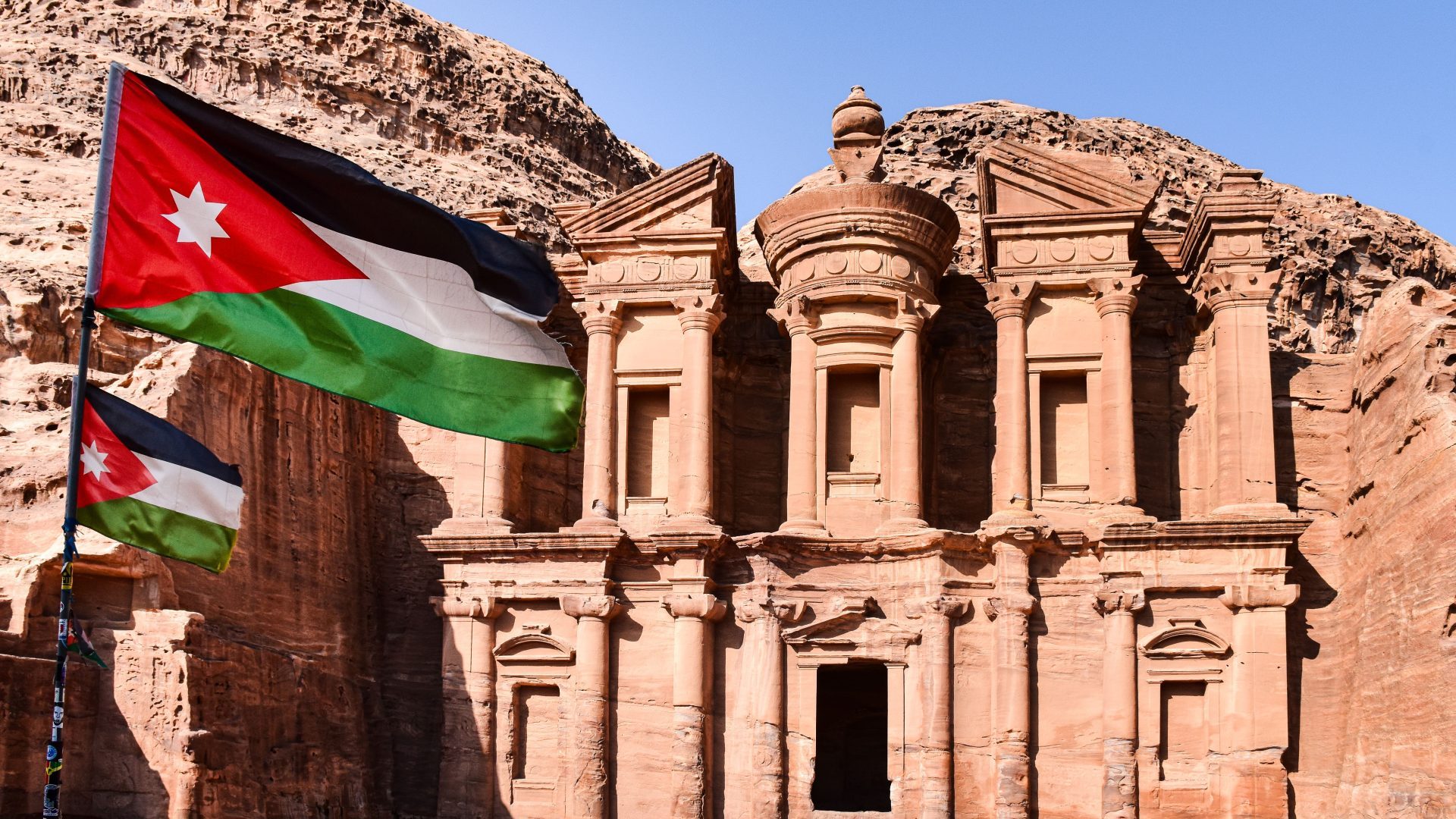 Ad Deir (The Monastery) in Petra, Jordan, with Jordanian flags in the foreground, monumental Nabataean rock-cut architecture surrounded by sandstone mountains under a clear blue sky