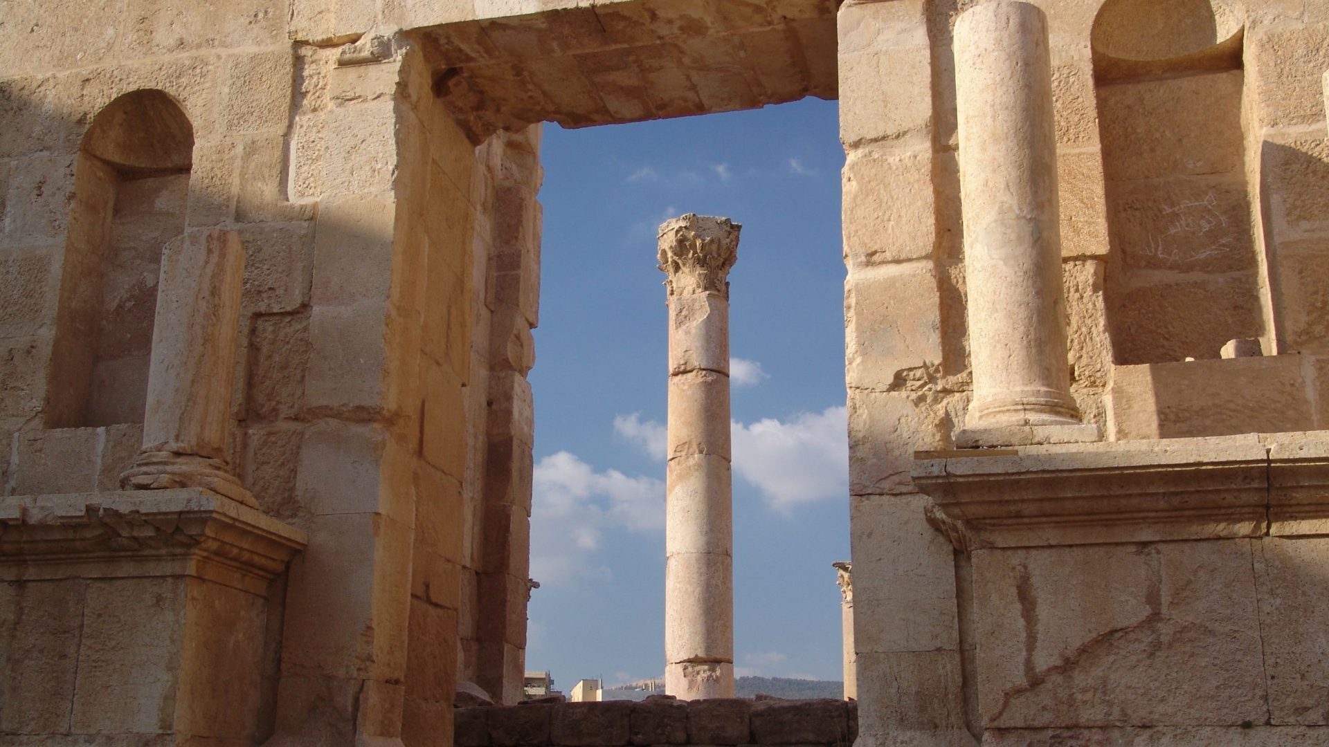 Ancient Roman column in Jerash, Jordan, framed by a historic stone doorway, featuring a Corinthian capital against a bright blue sky