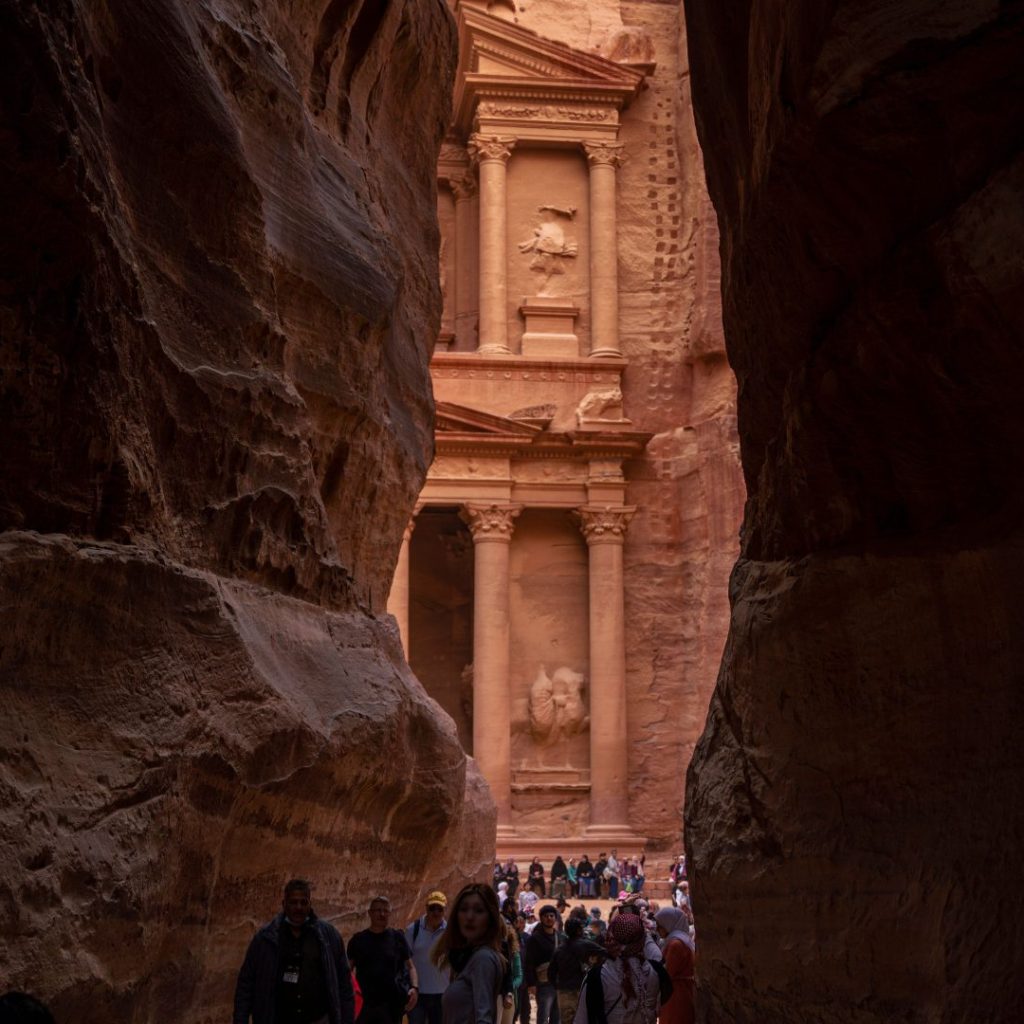 The Siq entrance view of Al-Khazneh (The Treasury) in Petra, Jordan, framed by narrow dramatic sandstone canyon walls with visitors walking toward the ancient Nabataean facade