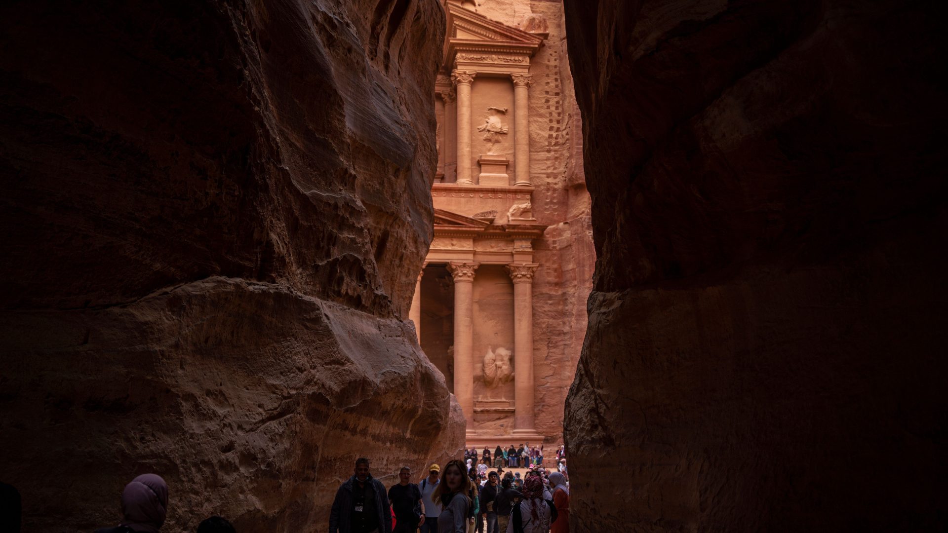 The Siq entrance view of Al-Khazneh (The Treasury) in Petra, Jordan, framed by narrow dramatic sandstone canyon walls with visitors walking toward the ancient Nabataean facade