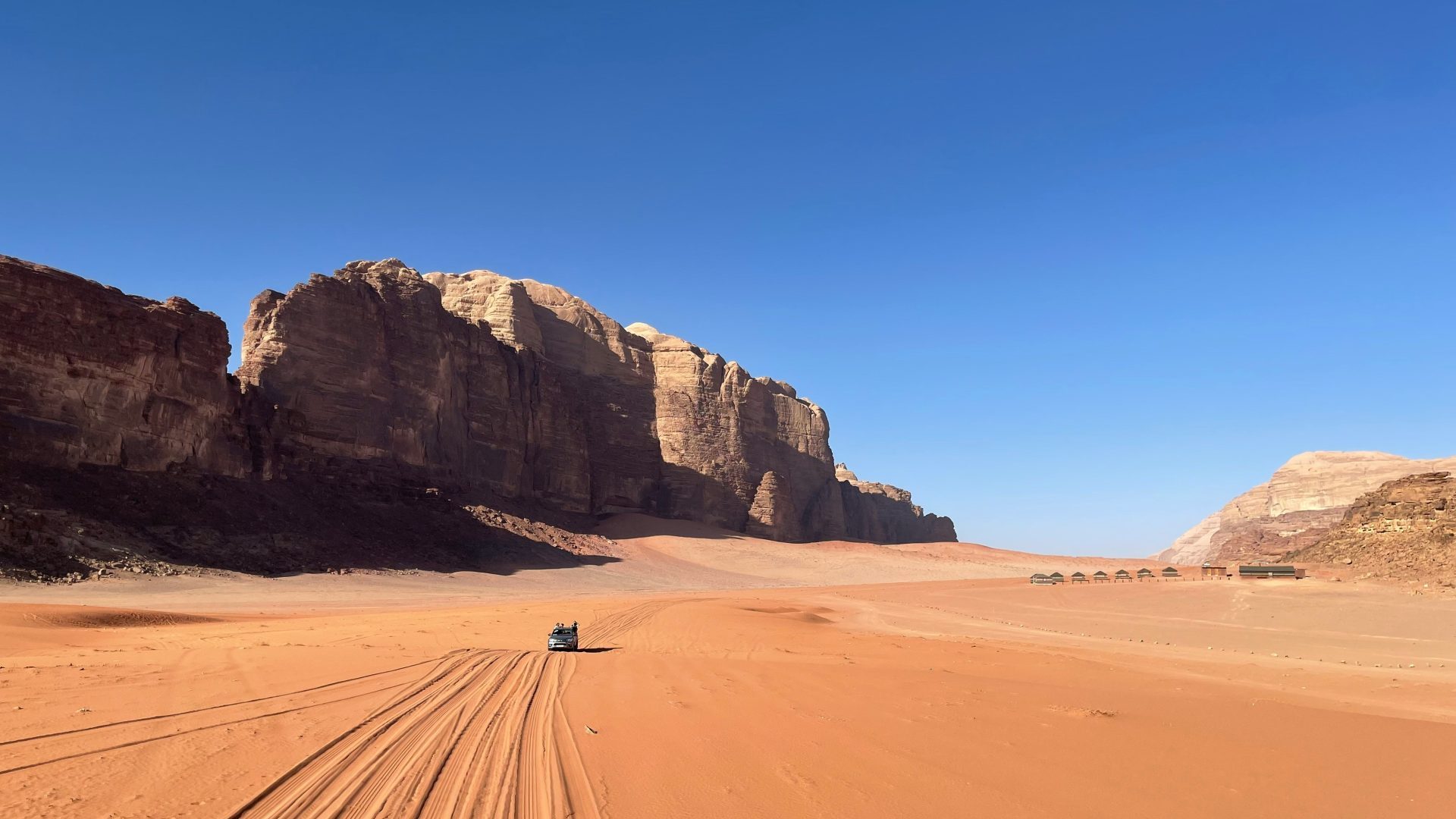Wadi Rum desert in Jordan, featuring vast red sand dunes, dramatic sandstone cliffs, and a lone 4x4 jeep crossing the desert under a clear blue sky