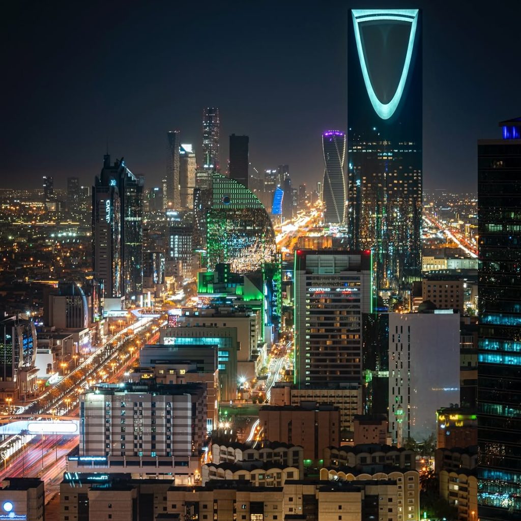 Riyadh skyline at night, featuring the turquoise-lit Kingdom Centre tower and brightly illuminated streets.