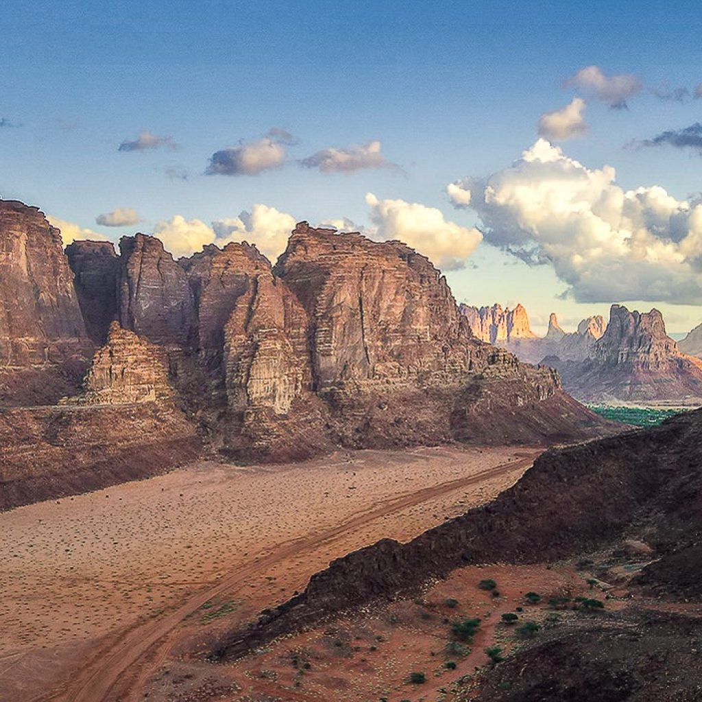 Wide desert valley with towering sandstone cliffs, a winding dirt track, and scattered greenery beneath a partly cloudy sky.