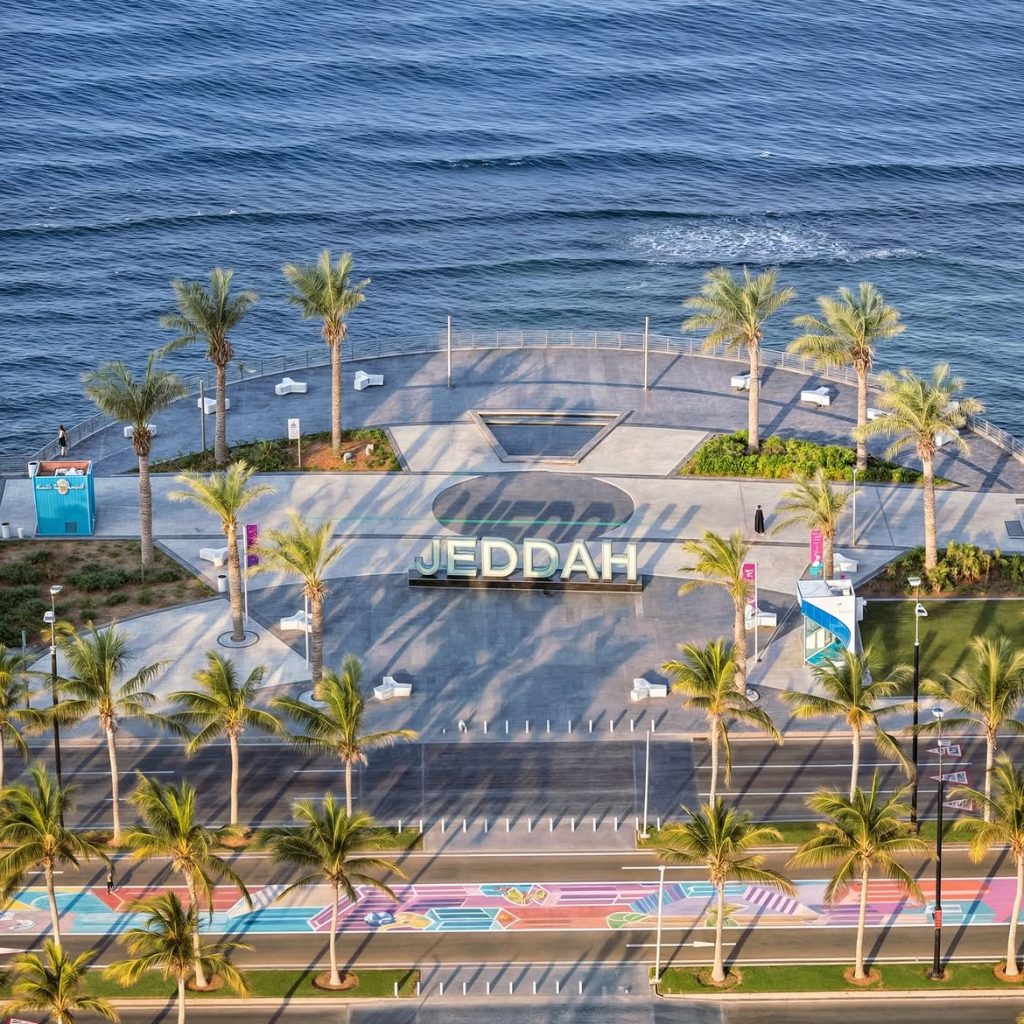 Aerial view of Jeddah’s waterfront with a large “JEDDAH” sign, palm trees, a colorful pathway, and the sea.