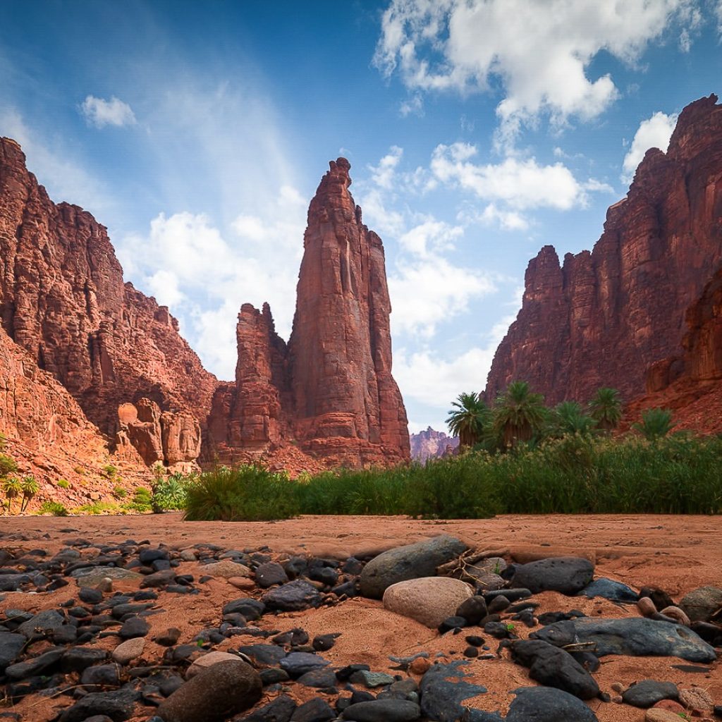 Red sandstone cliffs and a tall rock spire above a sandy, boulder-strewn valley with palm trees beneath a partly cloudy sky.