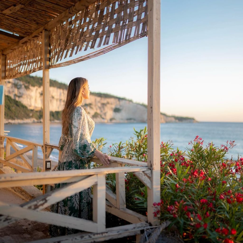 Woman standing on a shaded seaside terrace overlooking calm blue water and coastal cliffs, beside pink flowering plants.
