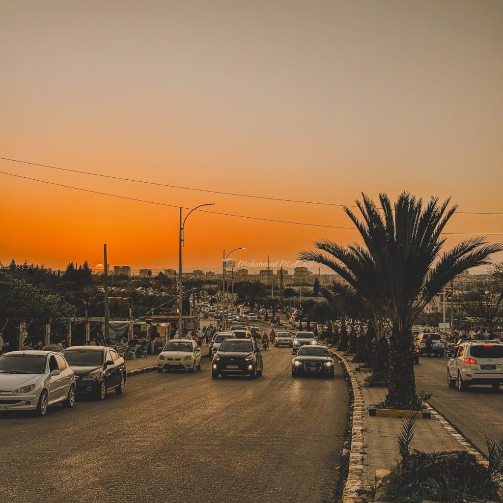 Cars traveling along a palm-lined city road beneath an orange sunset sky.
