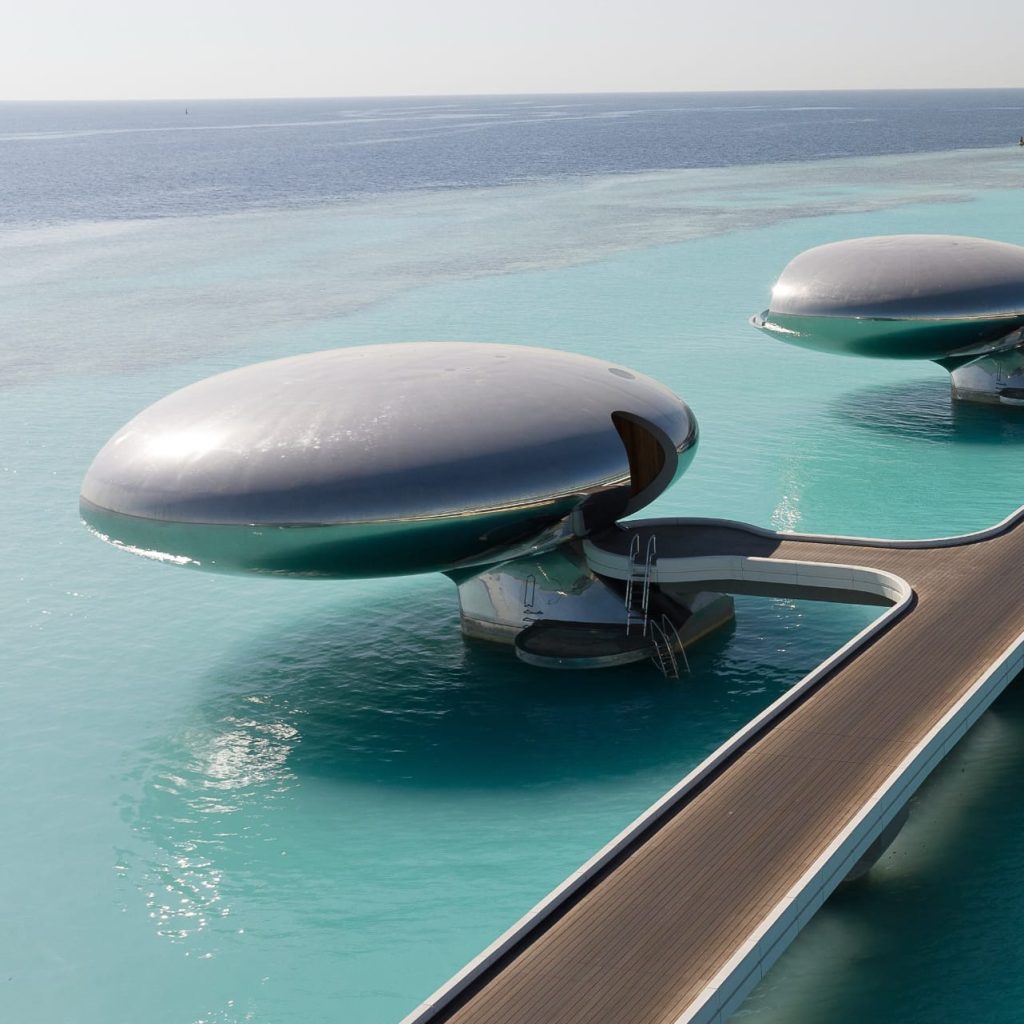 Silver dome-shaped overwater villas connected by a curved wooden boardwalk above clear turquoise water.