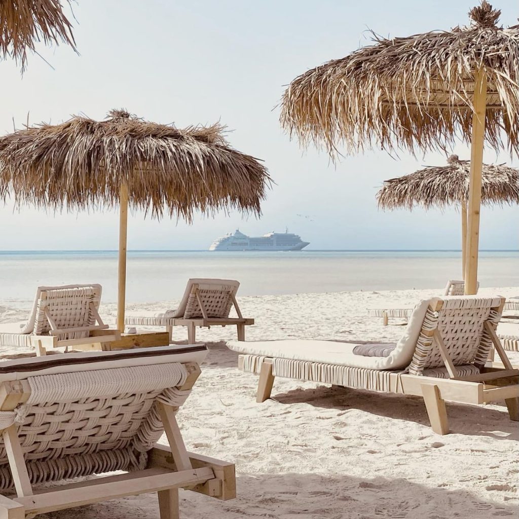 Wooden sun loungers beneath thatched umbrellas on a sandy beach, with calm sea and a cruise ship in the distance.