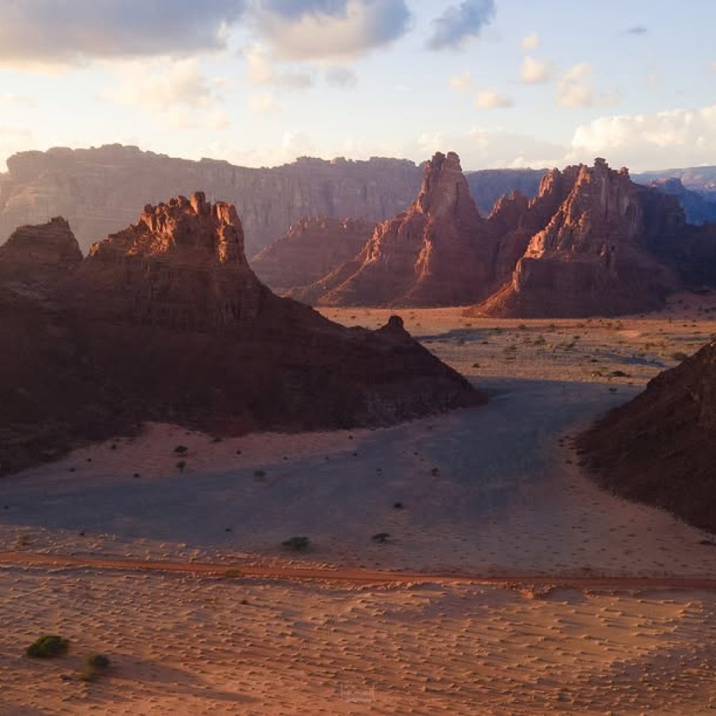 Rugged red sandstone formations rising above a sandy desert plain in warm evening light.