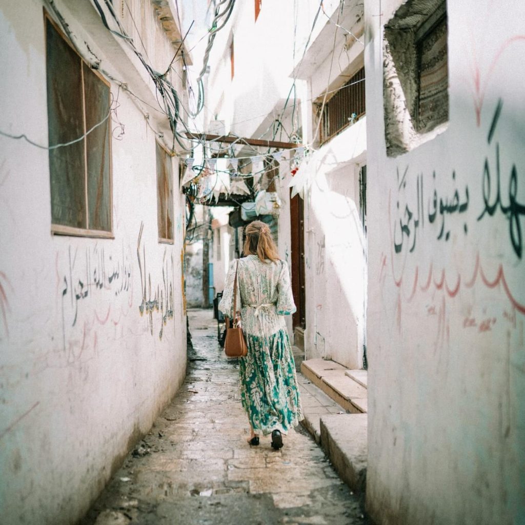 Woman in a green patterned dress walking through a narrow cobblestone alley with white walls, Arabic graffiti, and overhead wires.