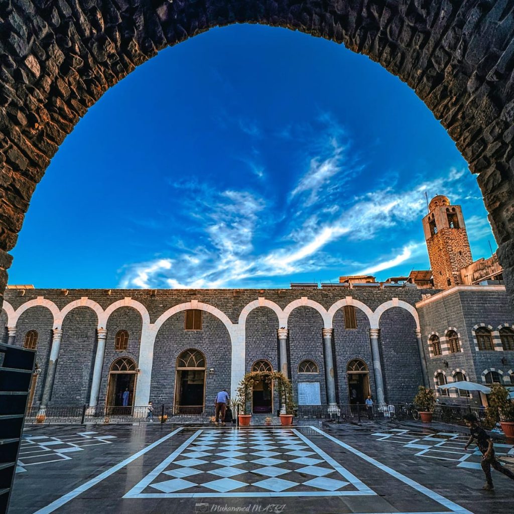 Stone courtyard with black-and-white patterned paving, arched colonnades, and a tower, viewed through a large stone arch.