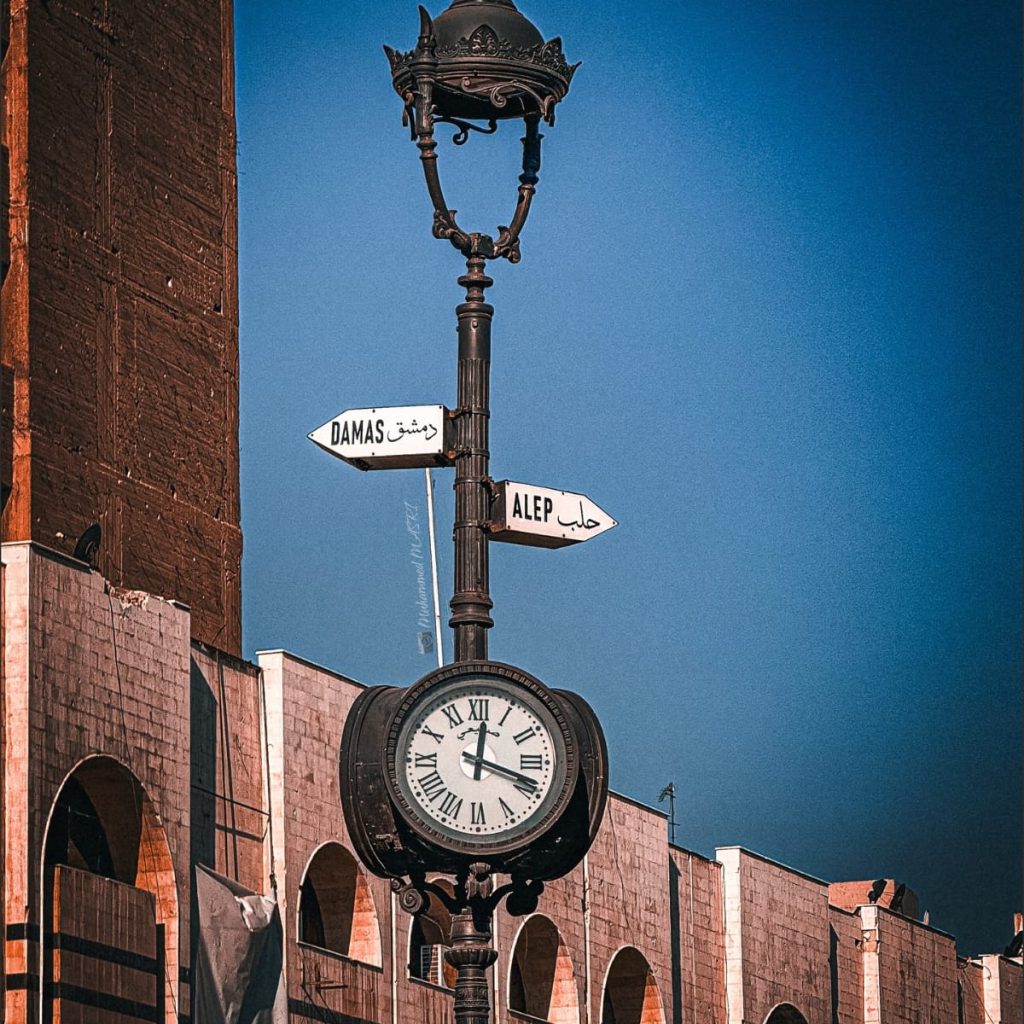 Ornate street clock and lamp post with signs pointing to Damascus and Aleppo, beside a stone building with arched windows.