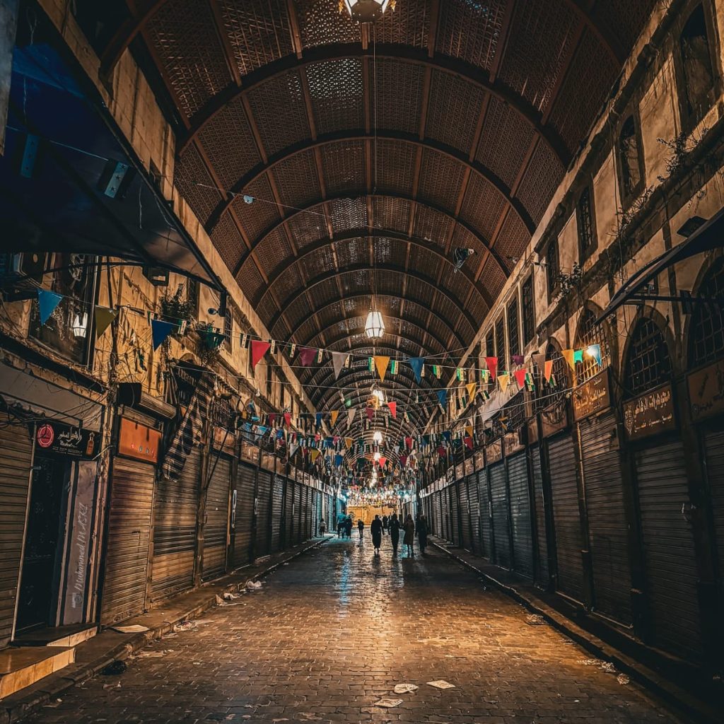 Covered market alley with shuttered shops, hanging lanterns, colorful bunting, and pedestrians on a cobblestone walkway.