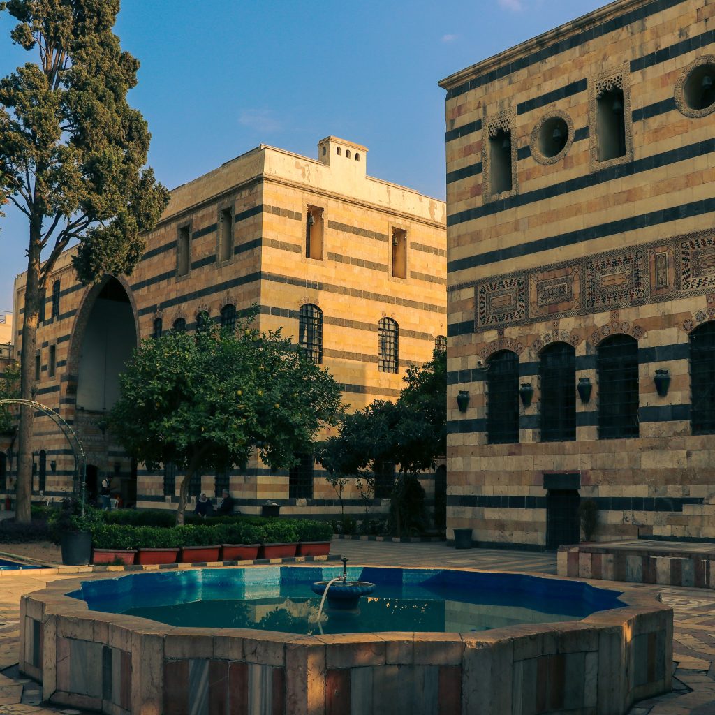 Historic courtyard with a blue-tiled fountain, leafy trees, and ornate buildings featuring alternating light and dark stonework.