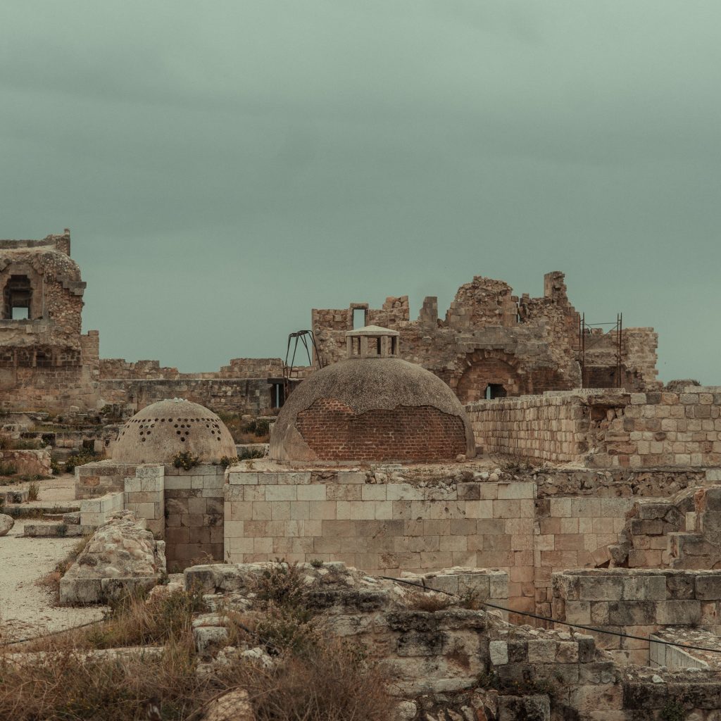Historic stone ruins with domed brick structures, weathered walls, and sparse vegetation beneath an overcast sky.